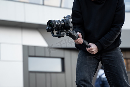 身穿黑色连帽衫的专业摄像师在三轴万向稳定器上手持专业相机。