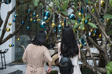 精美的蝴蝶摄影照片_两个女孩站在装饰精美的圣诞树旁，圣诞树上摆着蓝色和金色的玩具以及漂亮的蓝色蝴蝶玩具。
