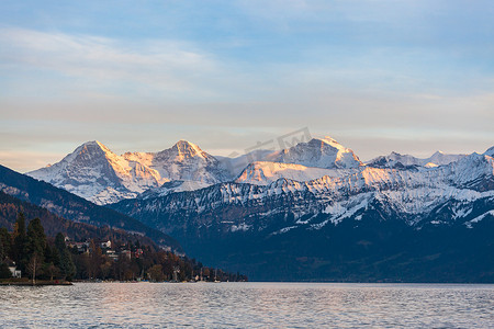 在阳光明媚的秋日，瑞士伯尔尼，从图恩湖 (Thunersee) 日落时，从图恩湖 (Thunersee) 欣赏著名的瑞士阿尔卑斯山峰在伯尔尼高地艾格峰北面、僧侣峰、少女峰的壮丽全景