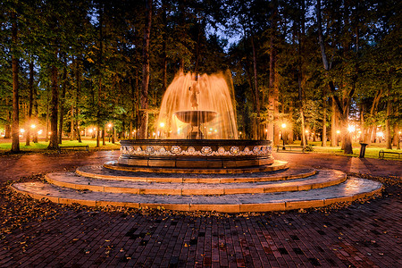 公园里的长椅摄影照片_夜间公园里有一个喷泉，水流模糊，灯笼照亮，铺着石头路面、树木和长椅。