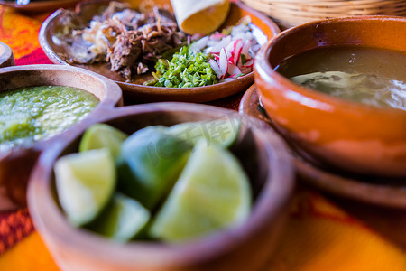 桌布墨西哥摄影照片_五颜六色的桌布上放着几碗酸橙片、青酱和羊肉汤