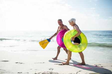 快乐的老年夫妇带着充气环和脚蹼在沙滩上散步