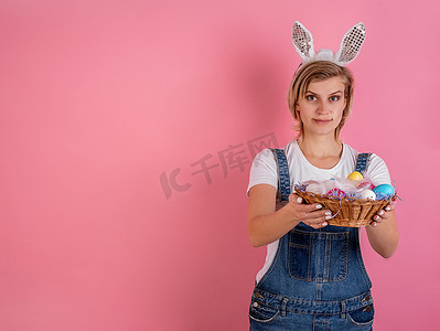 戴着兔子耳朵的年轻女子拿着篮子，篮子里有粉色背景中突显的彩色复活节彩蛋