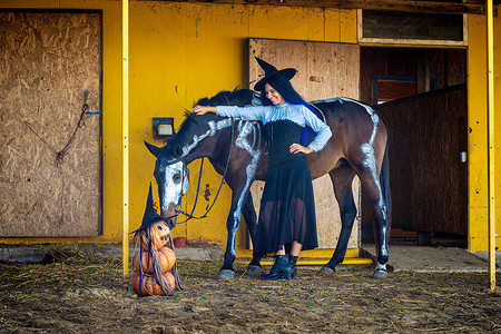 一匹马正在嗅一个即兴的南瓜雕像，一个打扮成女巫的女孩站在附近微笑