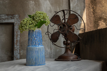 蓝色花束摄影照片_蓝色陶瓷花瓶中的绿色花束和阳台屋旧水泥墙的白色纹理桌布上的复古棕色金属风扇。