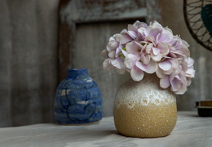一束白色的紫色花朵，黄色手工陶瓷花瓶，白色纹理桌布上的复古扇子，旧水泥墙。