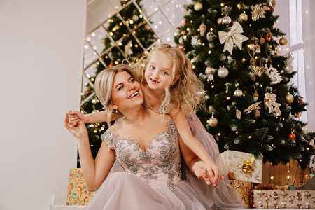 女儿抱着妈妈坐在圣诞树旁，拿着礼盒