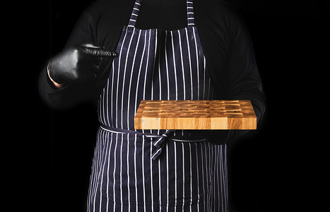 穿着蓝色条纹围裙和黑色衣服的男厨师站在黑色背景下，手里拿着一块长方形的木制厨房砧板
