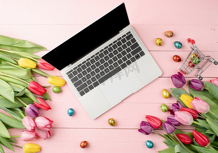 粉色木质背景上的笔记本电脑、彩色郁金香和复活节彩蛋的顶视图