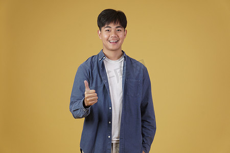 面带微笑、积极向上的亚洲年轻男子的肖像，穿着随意，大拇指竖起，赞同黄色背景中孤立的表情