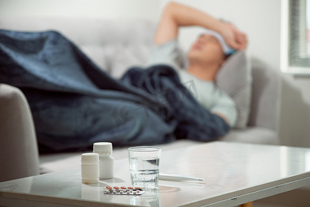 躺在沙发上的病态消瘦男子患有感冒和冬季流感病毒，在保健概念中有药片，在温度计上看温度