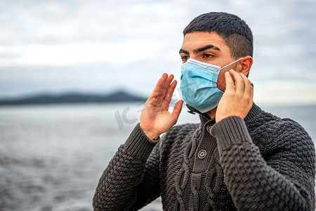 在海滨度假胜地户外穿着深色羊毛衫的帅哥刚刚戴上了防冠状病毒流行病毒的面部医疗防护面具 — 患有Covid-19社会疾病的年轻人在寒假