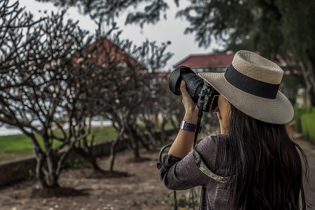 海边活动摄影照片_年轻女子戴着白色帽子，拿着相机拍摄海边的风景。