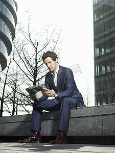穿着西装的年轻人坐在办公楼外的墙上看报纸