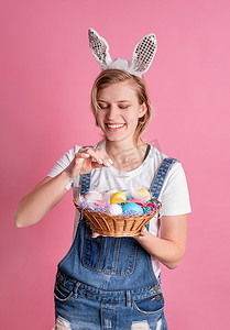 兔子粉色摄影照片_戴着兔子耳朵的年轻女子拿着篮子，篮子里有粉色背景中突显的彩色复活节彩蛋