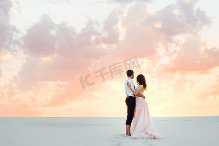 一对年轻夫妇，一个穿黑色马裤的男人和一个穿粉红色连衣裙的女孩正沿着白色沙滩散步