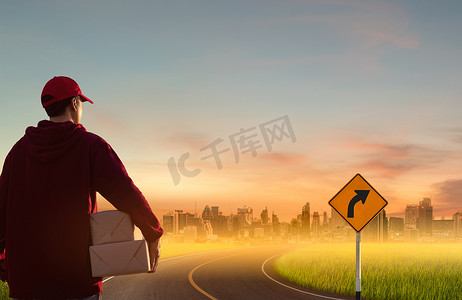 在具有城市背景的田野或草地上的自然日落时分，送货员在弯曲的道路上拿着纸箱。在线购物和快递概念。
