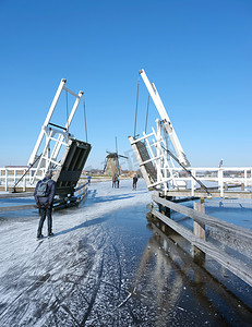 结冰的水摄影照片_荷兰阳光明媚的冬日，人们在运河结冰的水面上滑过 kinderdijk 的吊桥