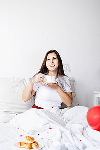 年轻的黑发女人坐在床上醒着，带着红心形气球和装饰品，喝咖啡吃羊角面包