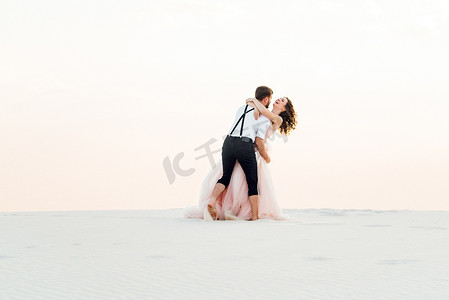 粉色沙滩摄影照片_一对年轻夫妇，一个穿黑色马裤的男人和一个穿粉红色连衣裙的女孩正沿着白色沙滩散步