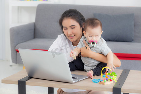 年轻的亚洲母亲在笔记本电脑上工作，同时在家照顾女儿，妈妈和女婴坐在一起使用笔记本视频通话、父母和亲密关系、室内、家庭和商业概念。