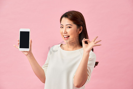 快乐的亚洲肖像美丽可爱的年轻女性兴奋地拿着手机并打着 ok 的手势，工作室拍摄在粉红色背景中被隔离