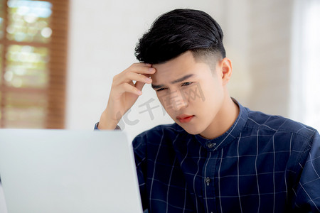 年轻的亚洲商人在笔记本电脑上工作时头痛，最后期限在家里的办公桌上，失败的商人筋疲力尽、疲倦、压力和担心、沮丧和失败。