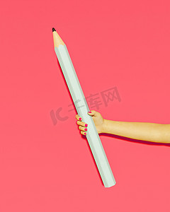 握着铅笔摄影照片_一只带粉红色指甲的女性手握着巨型铅笔