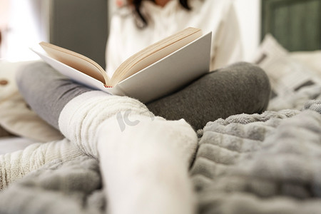 从低角度看，面目全非的女人穿着舒适的衣服坐在沙发上，放松地阅读一本前景模糊的白色羊毛袜的纸质书。