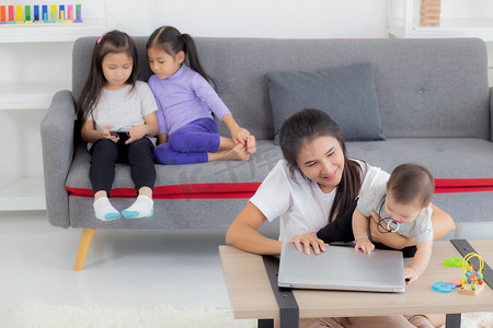 年轻的亚洲母亲在笔记本电脑上工作，同时在家照顾女儿，妈妈和女婴坐在一起使用笔记本视频通话、父母和亲密关系、室内、家庭和商业概念。