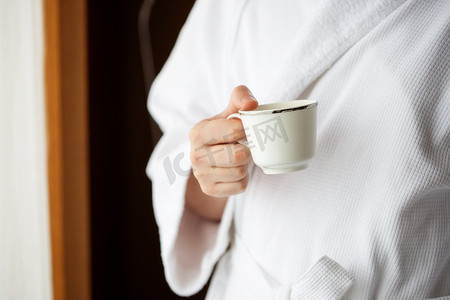 一位身穿白色毛巾布晨衣的男士在窗前拿着咖啡杯。