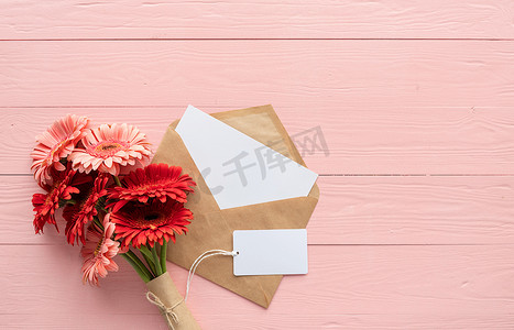 粉色木桌上的红色大丁草雏菊花、信封和空白标签