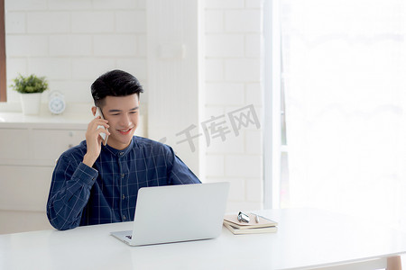 年轻的亚洲男子通过笔记本电脑在家打电话和工作，自由职业者使用笔记本电脑上网并谈论智能手机、家庭生活、商业和沟通理念。
