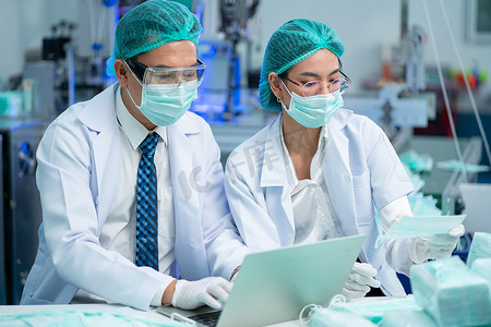 质量控制部分的两名工厂工人男女使用笔记本电脑在工厂机器前讨论口罩产品。