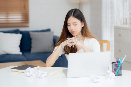使用笔记本电脑工作的年轻亚洲女性认为创意项目和纸在家里的桌子上皱巴巴的有问题，女孩使用笔记本带着沮丧和麻烦，商业和自由职业者的概念。
