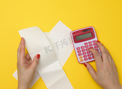 手帐背景摄影照片_两只女性手拿着纸质支票和黄色背景上的粉红色计算器。