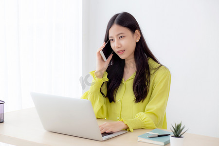 年轻的亚洲女商人在家庭办公室工作并打电话在家里取得成功，女商人在办公桌上使用笔记本电脑，女性在工作场所使用智能手机说话，沟通理念。