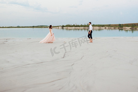 粉色沙滩天空摄影照片_一对年轻夫妇，一个穿黑色马裤的男人和一个穿粉红色连衣裙的女孩正沿着白色沙滩散步
