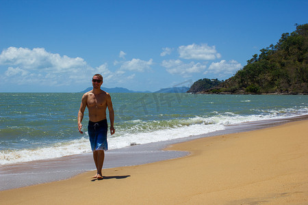 肌肉发达的白人男子走在沙滩上，当海浪在他身后拍打时微笑，苦难角，澳大利亚昆士兰