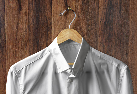 一件白色礼服衬衫的特写镜头挂在一个木制衣架上，衣架挂在深色木镶板墙上的挂钩上。