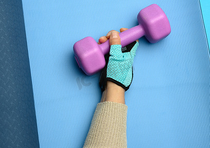 蓝色运动手套中的女性手握着蓝色背景上的紫色一公斤哑铃，