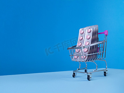 蓝色背景下超市手推车中的粉色药片起泡，带有倾斜的时尚天际线和复制空间。