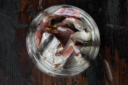 小木桌摄影照片_腌制的咸凤尾鱼片，在玻璃罐中，在旧的深色木桌背景上。顶视图平躺