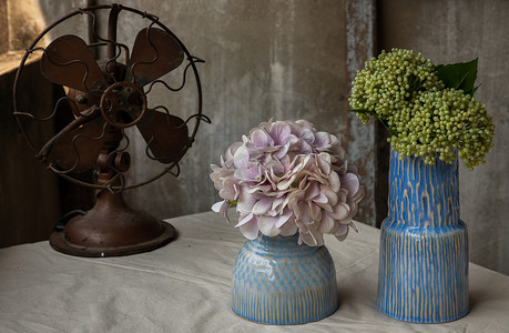 两个蓝色手工陶瓷花瓶和老式棕色金属风扇中的花束，白色纹理桌布上有旧水泥墙，位于阳台屋。
