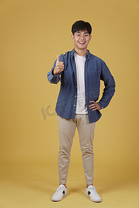 好帅的表情摄影照片_面带微笑、积极向上的亚洲年轻男子的肖像，穿着随意，大拇指竖起，赞同黄色背景中孤立的表情