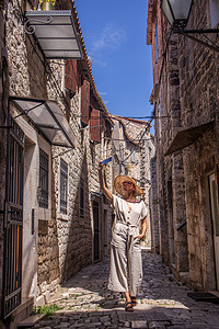 美丽的金发年轻女性旅行者戴着草帽太阳帽，在克罗地亚亚得里亚海的一个古老的传统沿海小镇观光和享受暑假时自拍。