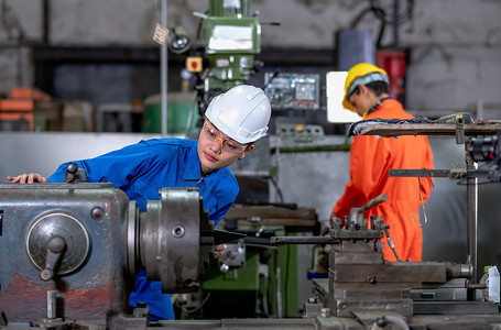工厂其他摄影照片_亚洲工厂女工看着机器的一部分并检查功能，而她的同事在后台与其他机器一起工作。
