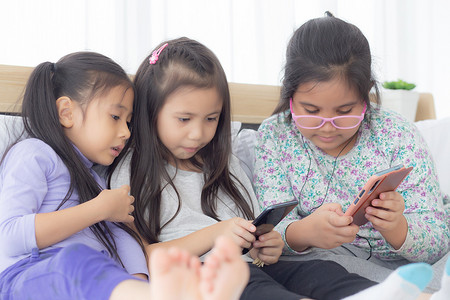 正能量手机海报摄影照片_快乐的亚洲小孩和朋友在家里的沙发上玩智能手机，孩子们在沙发上一起使用手机，女孩看智能手机以获得娱乐、生活方式和沟通理念。