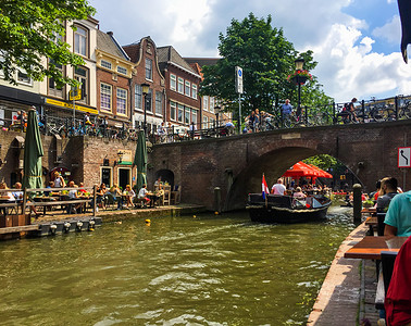 2018年戊戌摄影照片_乌得勒支/荷兰-2018 年 6 月 3 日：美丽建筑的壮丽景色、一座带自行车的桥、一艘旅游船和许多街边咖啡馆，在 Oudegracht（旧运河）附近有让人放松的人