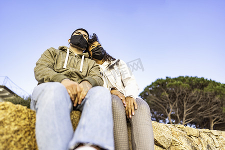 年轻的多种族情侣坐在墙上，戴着黑色安全防护面罩，头对头看着日落时的地平线。